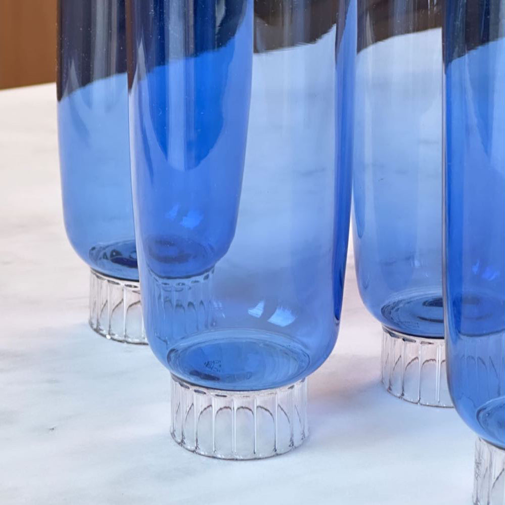 The image offers a detailed view of colored champagne glasses, highlighting their exquisite cobalt blue colored glass. The lower section of each flute displays a beautiful stemmed bottom, intricately designed with a ribbed texture that captures and bends the light, adding a touch of elegance and visual interest. The flutes transition from this ornate base into a striking cobalt blue color, which gives these colored champagne glasses a vibrant and luxurious appearance.