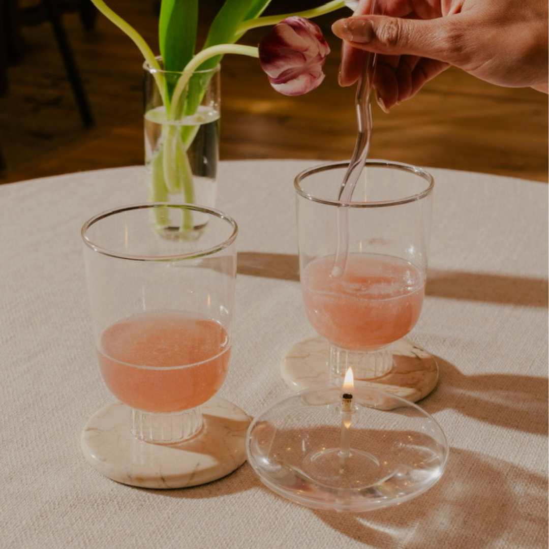 Set of two stemless wine glasses with rose sprinkled with glitter. The stemless wine glass sits on a marble coaster. Made of 100% borosilicate glass it is lightweight yet durable and dishwasher safe. The clear champagne glasses has been used as bud vase with slender tulips. This scene suggests a relaxed yet sophisticated setting, possibly a romantic dinner or a cute dinner with the girlies.