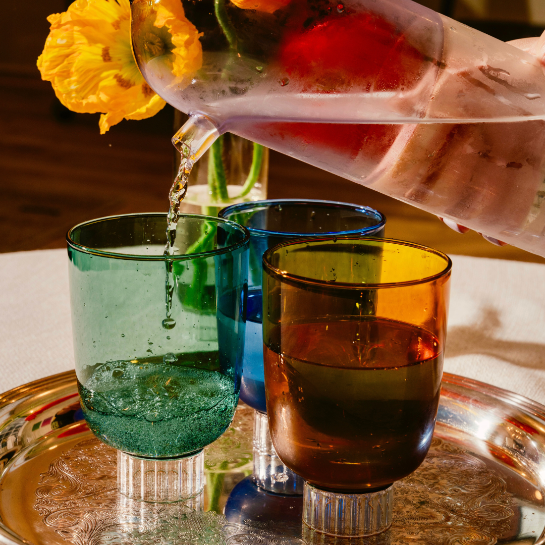 Discover Sprezz's premium colored glassware, water is being poured from a color blocked lilac water carafe into a teal colored glass from a stemless wine set. The image features a silver tray with a set of Sprezz's Romantic vintage-style glasses in rich hues of forest green, cobalt blue, and amber.