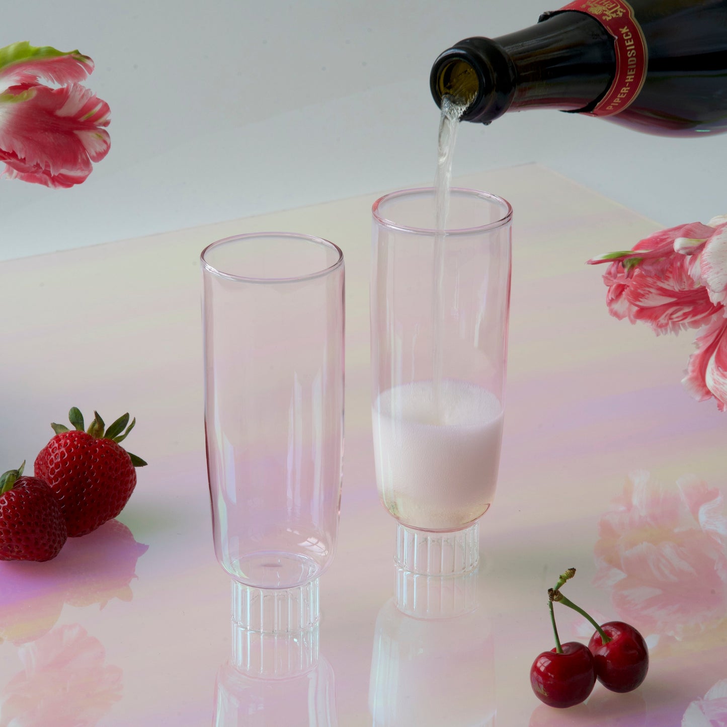 Contemporary champagne flutes being filled with sparkling pink champagne in front of a iridescent pink background with strawberries and cherries, showcasing Sprezz's unique and colored champagne glasses, ideal for a brunch party or a spring tablescape.