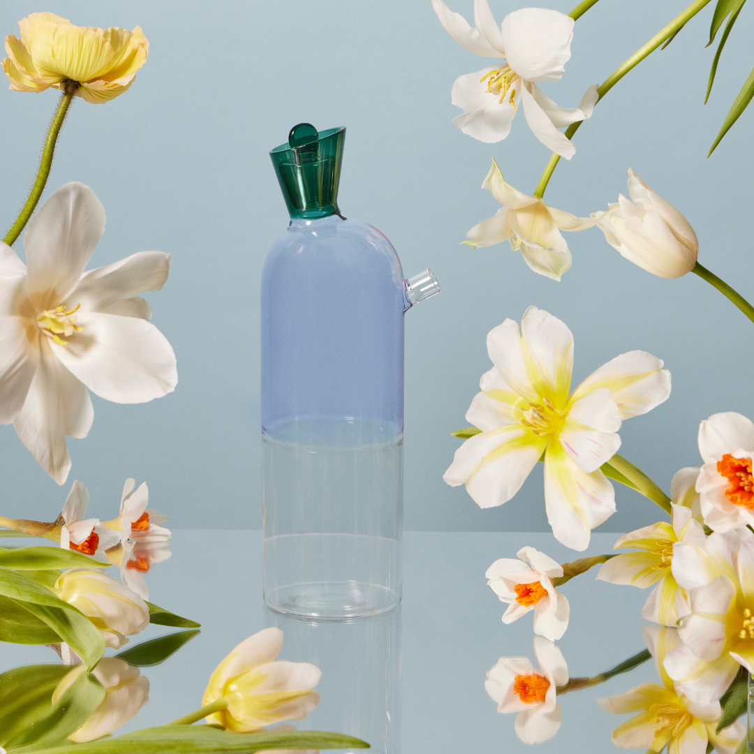 A whimsical lilac carafe and pitcher with a tapered body and anemerald green stopper, on a mirrored table. The decanter is surrounded by beautiful creamy white and soft yellow flowers, adding a touch of whimsical elegance the dinner table.